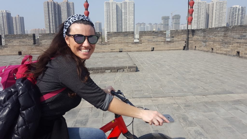 Mariacristina smiling and cycling, with the edge of Xi'an city wall in the background
