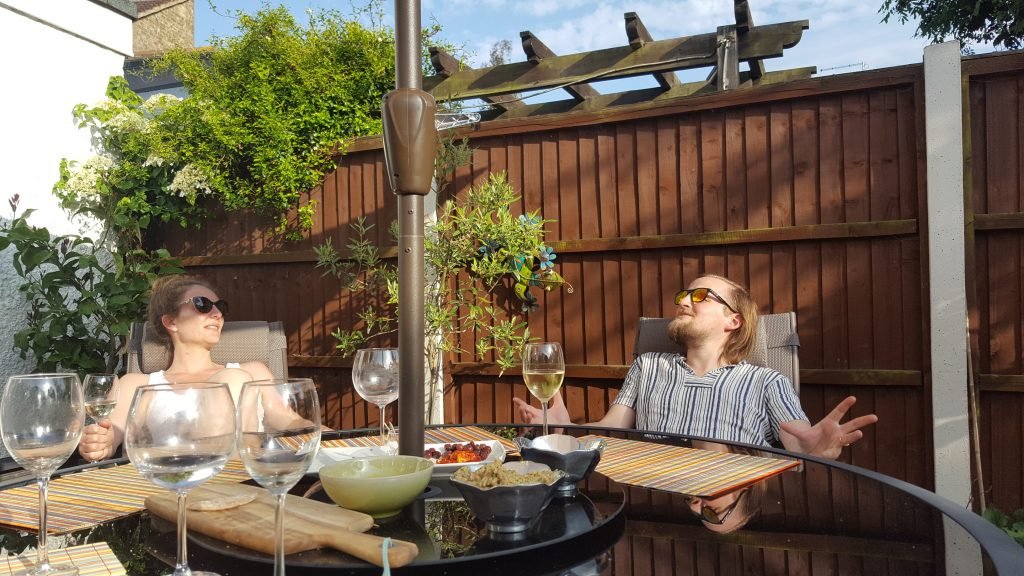 Rae and George looking happy sitting at a garden table with wine glasses and sunglasses