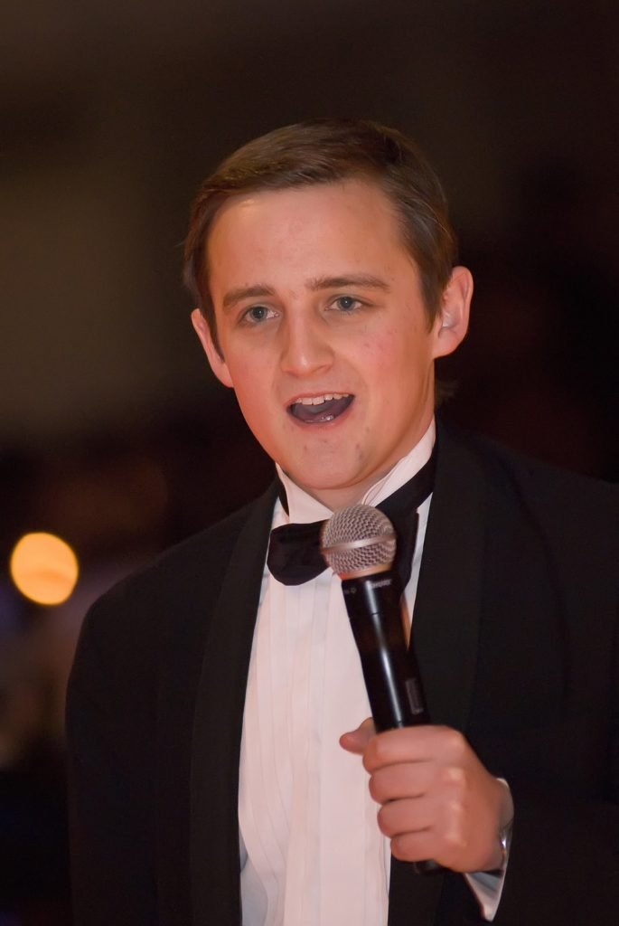 George with no beard, looking younger, but wearing black tie and talking into a microphone