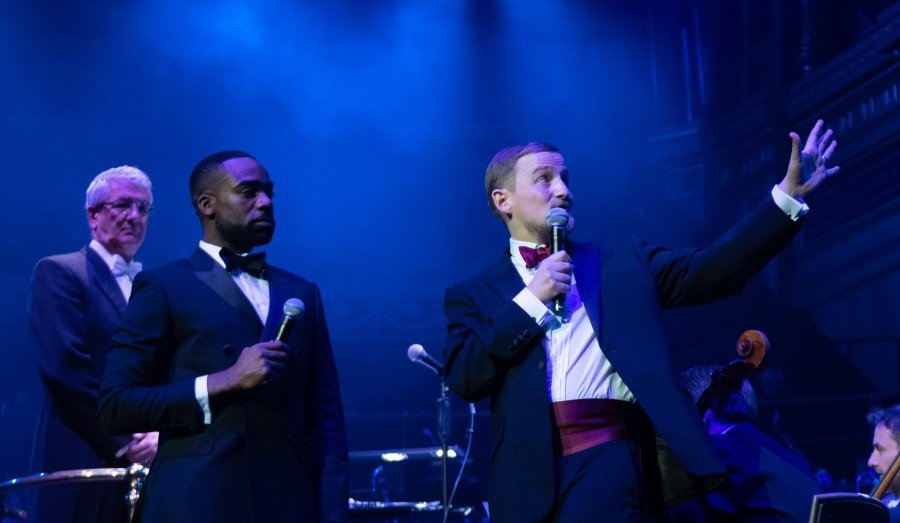 George and Ore Oduba onstage in black tie, with George waving his left arm up towards the choir and speaking into the microphone